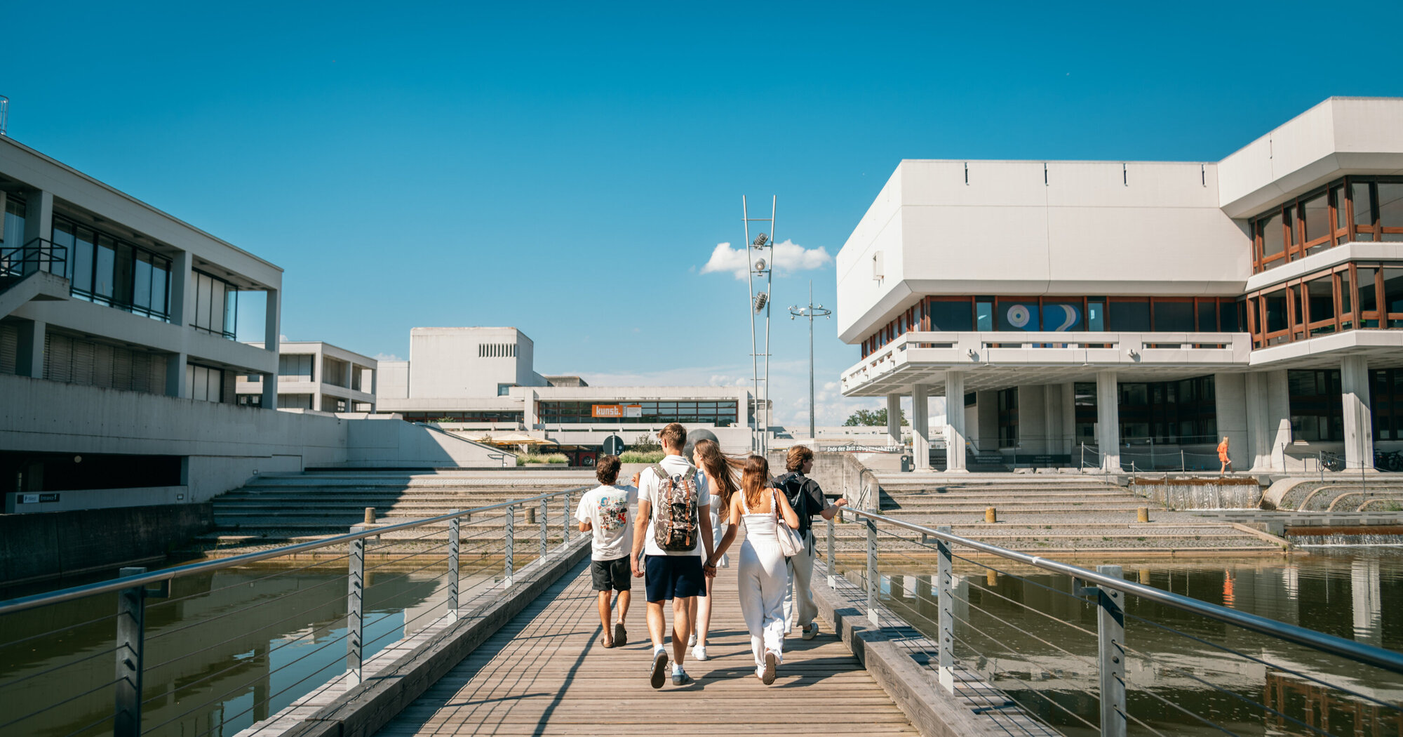 Studierende gehen auf der Brücke über den Uni-See in Richtung Zentrales Hörsaalgebäude der Universität Regensburg.