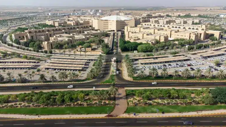صورة لجامعة القصيم تُظهر الحرم الجامعي الحديث بمبانيه البيجة، وأشجار النخيل والمساحات الخضراء التي تتوزع وسط البيئة الصحراوية المحيطة وتبرز في الصورة مواقف السيارات والطرق التي يسير عليها عدد من المركبات تظهر الكلية التطبيقية وبرامج الدبلوم عن بُعد وإمكانية التقديم المفتوح، في مشهد يعكس مشهد الحداثة والتنظيم مع التأكيد على الالتزام بالزي الرسمي مثل الشماغ أو الغترة.