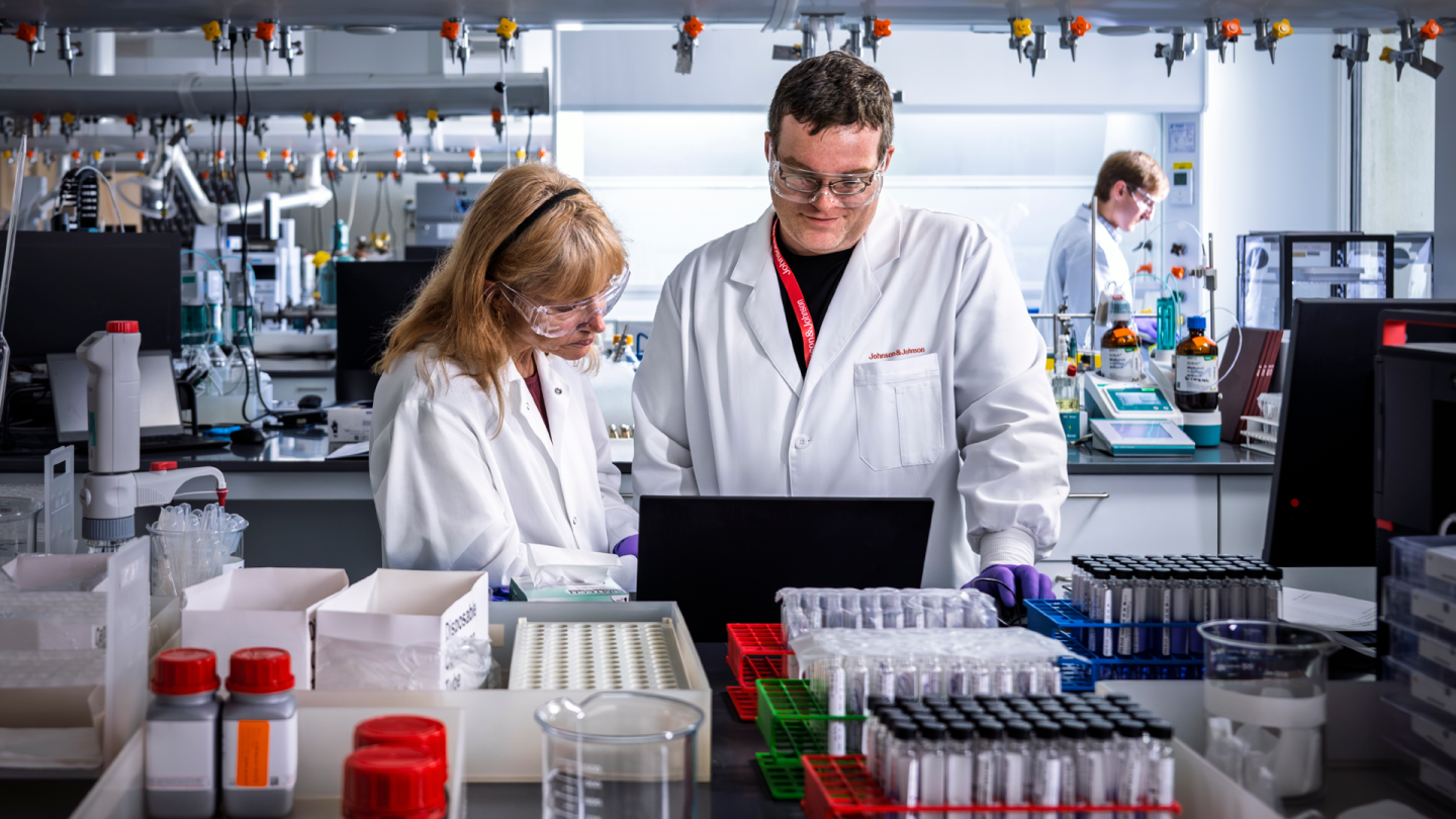 Researchers in a laboratory reviewing data on a laptop while working with test samples and scientific equipment.
