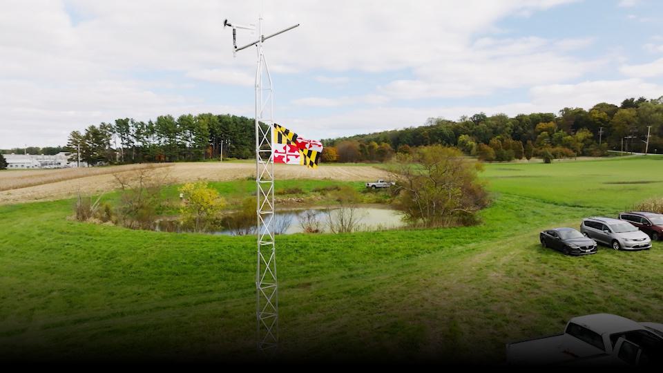 Maryland flag waving in a field with a pond. Cars parked on field.