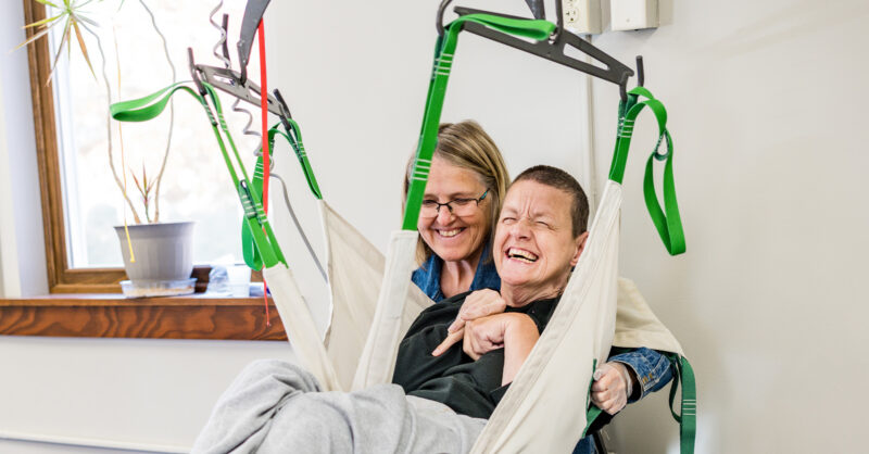 A DakotAbilities direct support professional smiles warmly while assisting a joyful adult with a physical disability using a ceiling track lift sling system. The client laughs during the transfer process, reflecting the compassionate, person-centered care provided at DakotAbilities in Sioux Falls, South Dakota. An adaptive power wheelchair is visible in the foreground, highlighting the organization's commitment to empowering adults with physical and developmental disabilities through dignified, skilled residential and habilitation support services.