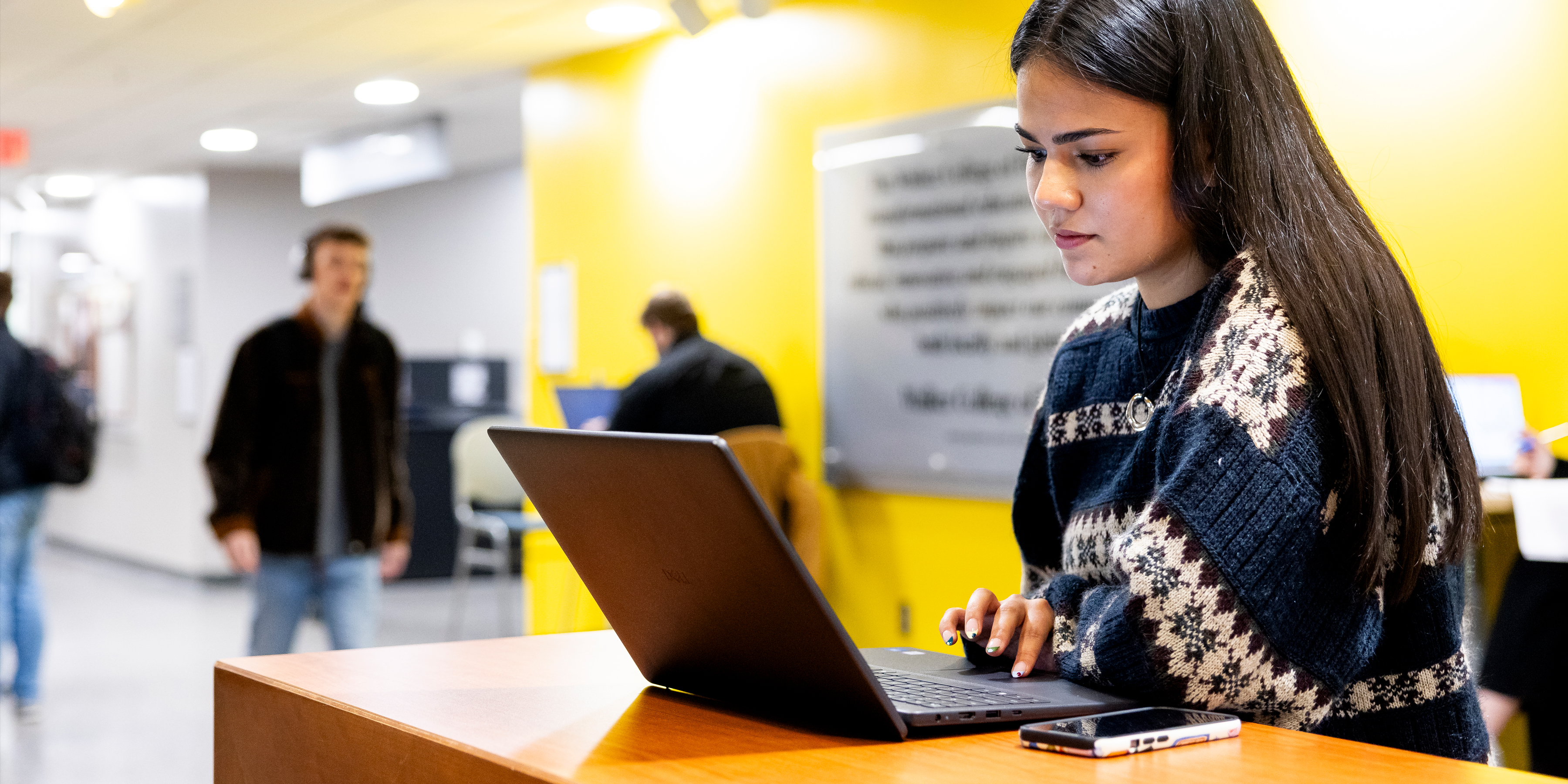 Person working on a laptop at a desk in a brightly lit indoor space with other people in the background.