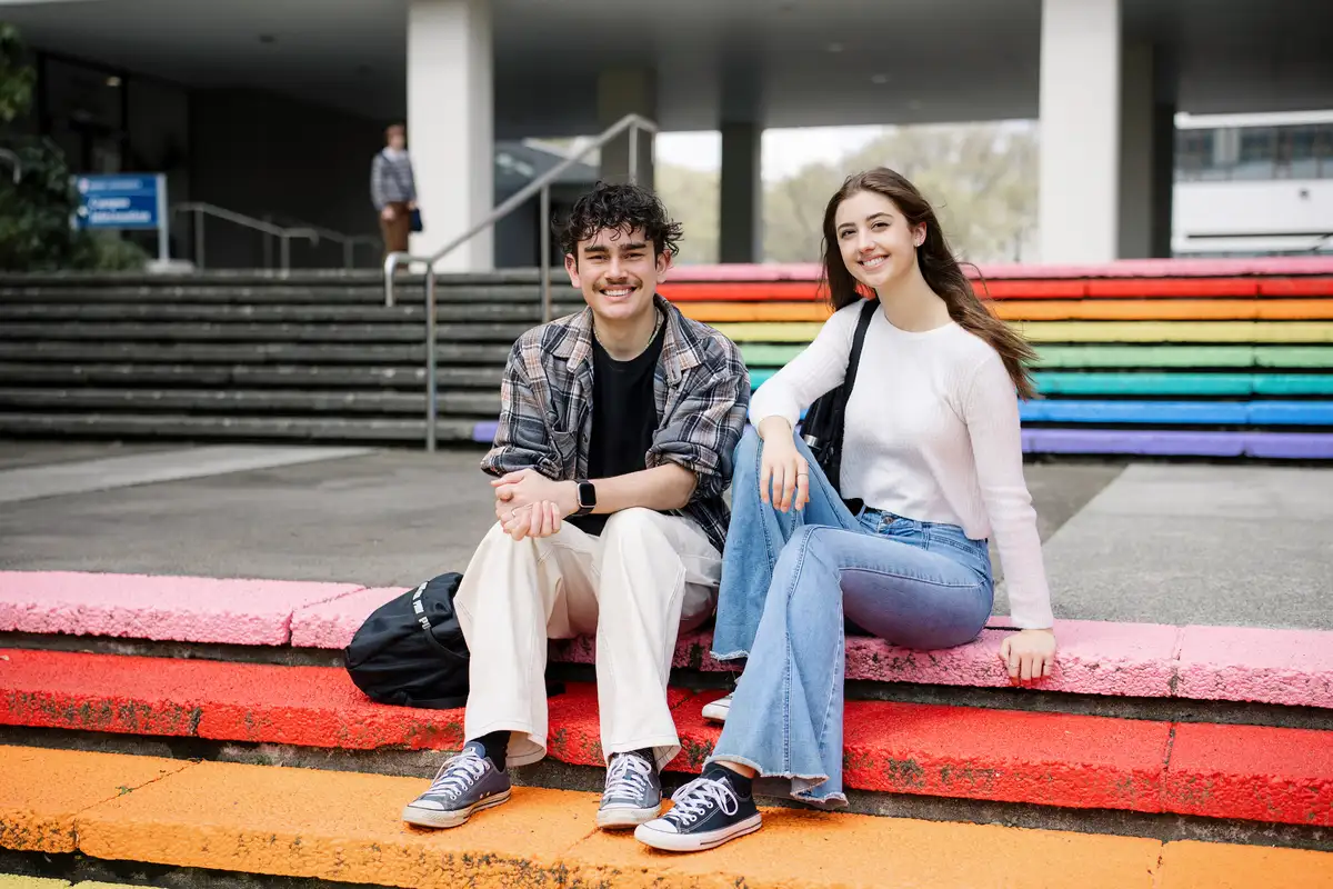 Two students sitting on rainbow painted steps outside Massey University campus.